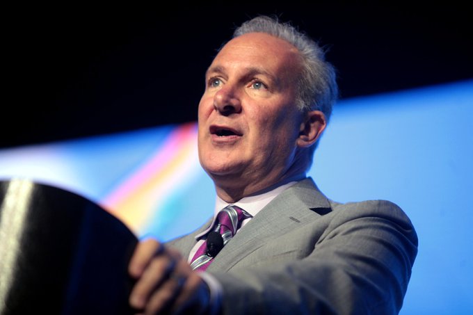 A middle-aged man in a gray suit and striped tie stands at a podium, appearing to give a speech or presentation against a dark backdrop with blue lighting. The image relates to the accompanying post about Peter Schiff and his views on tokenized gold, suggesting the speaker is Peter Schiff himself, known for his promotion of gold investments and criticism of Bitcoin.