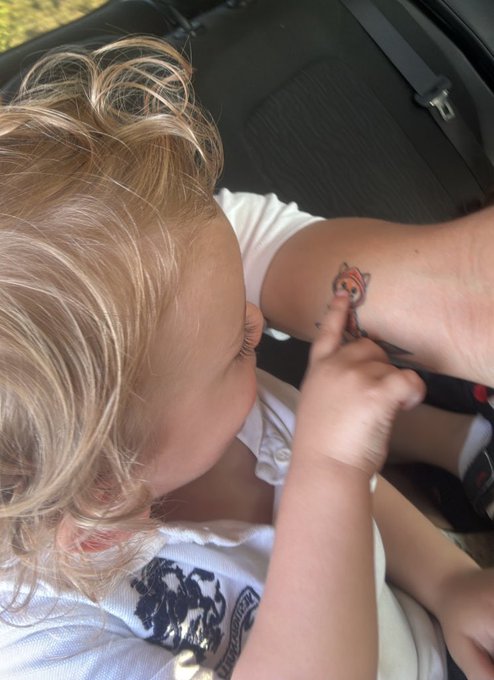 A young child with blonde hair, wearing a white shirt, looks closely at a small tiger tattoo on an adult's arm.