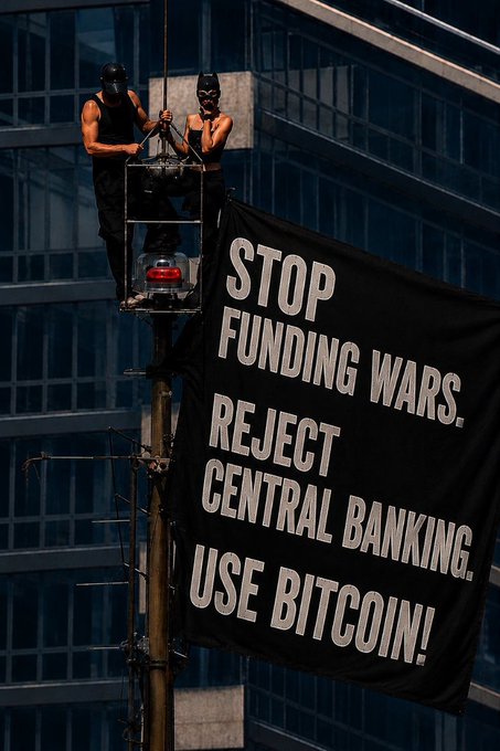 A photo of two masked activists on a pole-mounted platform unfurling a large black banner reading 'STOP FUNDING WARS. REJECT CENTRAL BANKING. USE BITCOIN!' in front of a glass office building.