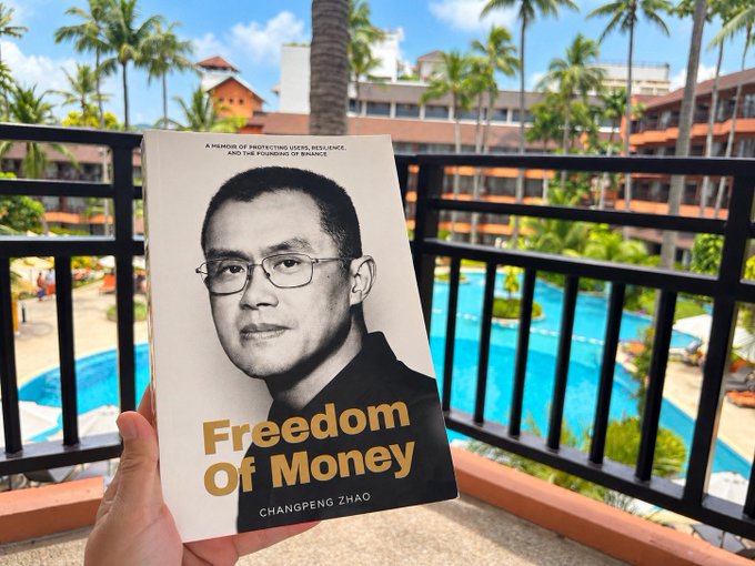 A hand holding the book 'Freedom Of Money' by Changpeng Zhao, with subtitle 'A MEMOIR OF PROTECTING USERS, RESILIENCE, AND THE FOUNDING OF BINANCE', photographed against a tropical resort pool background.
