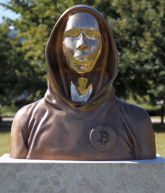A bronze-colored statue of a hooded figure with a highly reflective golden face, featuring a Bitcoin symbol on its chest, set against a background of green trees.