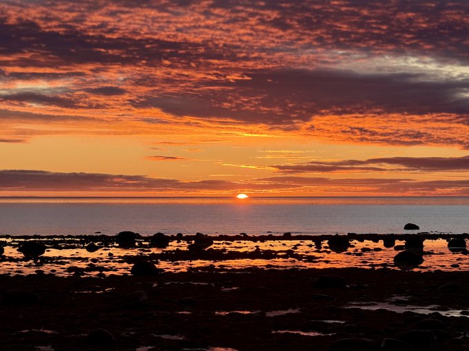 A scenic view of a sunset over the ocean, with a vibrant orange sky filled with scattered clouds above a rocky shoreline.