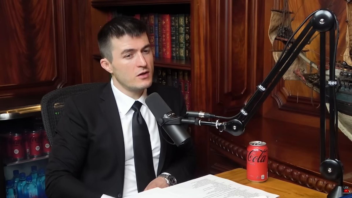 The image features a man dressed in a suit, sitting at a desk with a microphone in front of him. The man appears to be talking into the microphone or giving an interview. The background shows a bookshelf filled with numerous books, suggesting that he might have been discussing business or technology topics. A Coca-Cola can is also visible on the desk, indicating that this could be related to a discussion about the company's history and its impact on society.