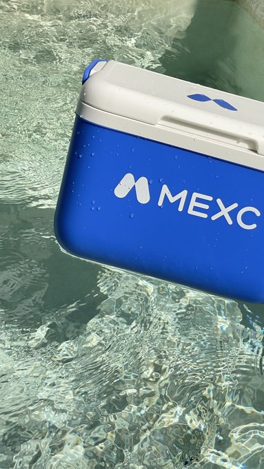 A blue and white MEXC branded cooler floating in clear, shallow water.