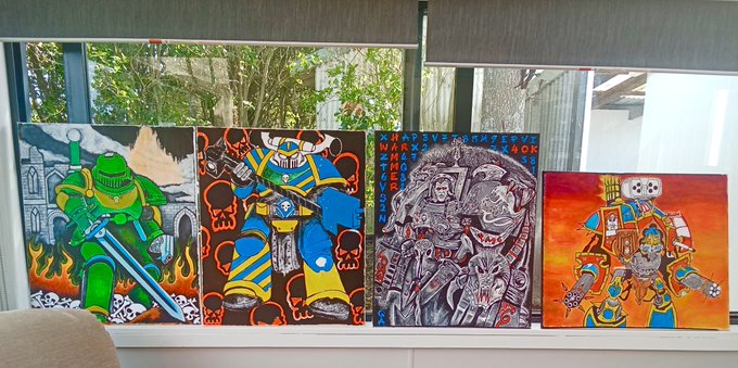 Four Warhammer-themed paintings displayed on a windowsill, featuring various Space Marine chapters in green, blue, grey, and orange color schemes.