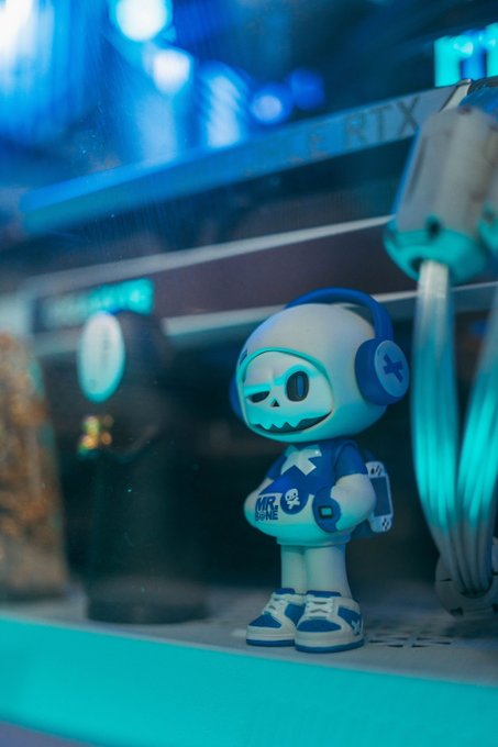 A small white and blue collectible figure wearing headphones stands in front of a computer setup bathed in cyan lighting, with an RTX graphics card visible in the background.