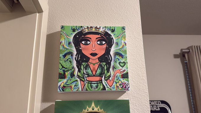 A small square canvas painting of a green-clothed girl with a crown holding a heart, signed 'CSG Art', displayed on a white shelf.