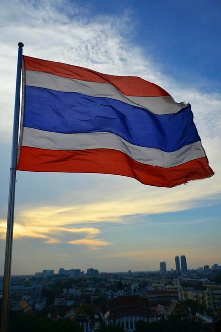 The Thai national flag waves against a cloudy sky, with a city skyline visible in the background. The image relates to the post as it visually represents Thailand, the country set to implement stricter regulations on large USDT transactions and cash deposits exceeding $150,000.