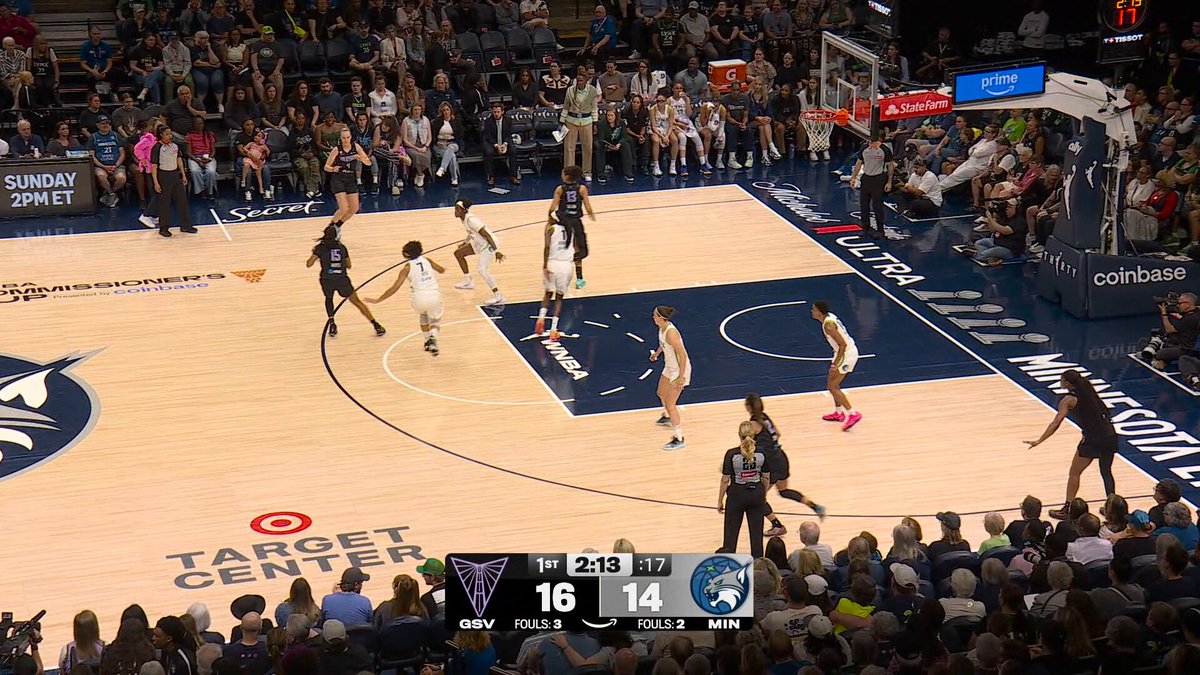 The image shows a basketball game with players on the court, including one player holding a basketball. The crowd of people watching the game can be seen in the background. There are also two TVs visible in the scene, possibly displaying the game or related content. The main focus is on the basketball game and its participants, while the surrounding elements provide context to the image's location and purpose.