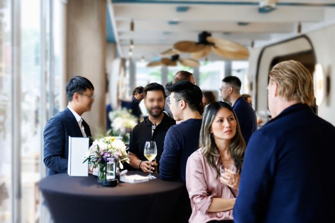 Attendees network at a well-attended Singapore event, with men in suits and a woman in a pink blouse engaging in conversation over wine and floral arrangements, capturing the bustling atmosphere of the speaker's action-packed week featuring 16 events and meetings with 400+ industry participants.