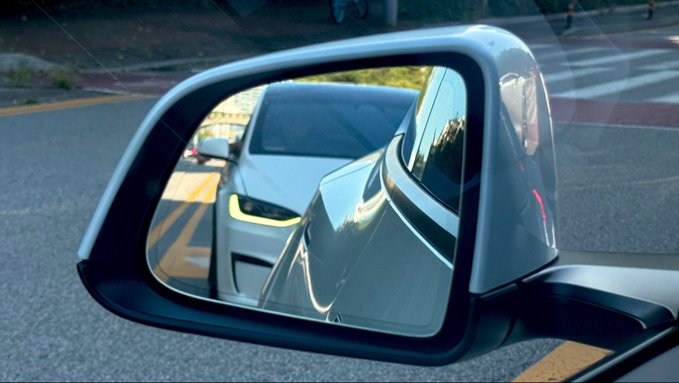 A view of a car's side mirror reflecting the angular front end of another gray vehicle, possibly a Tesla Cybertruck, stopped at an intersection.