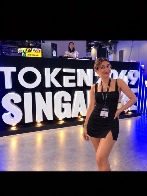 A photo of a woman in a black dress standing in front of a 'TOKEN2049 SINGAPORE' sign, wearing a lanyard and posing with her hand on her hip.
