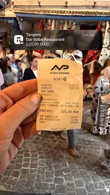 A person holds a receipt from Attijari Payment for 'DAR BABA' restaurant showing a charge of 520.00 MAD, with a mobile notification showing 'Tangem - Dar Baba Restaurant - 520,00 MAD - 29m ago' overlaid on a bustling market scene.