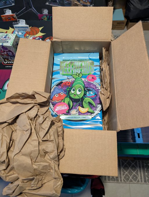An open cardboard shipping box filled with brown packing paper, revealing a 'Veggie Madness' limited edition comic book featuring a green character and the label 'Shells Kitchen'.