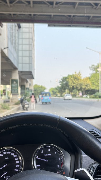 A view from inside a car showing the steering wheel and dashboard speedometer, looking out through the windshield at a sunlit street with a bus, motorcycle, and pedestrians.
