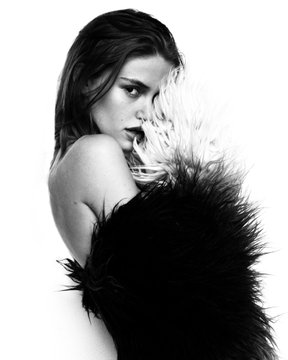 Black and white portrait of a woman with dark hair, wrapped in a dark, feathery garment, looking over her shoulder at the camera.