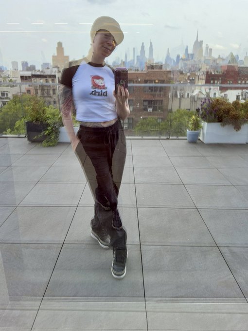 A photograph of a person on a tiled rooftop taking a mirror selfie, wearing a bucket hat, a white t-shirt with a red logo reading "child", and dark pants, with a city skyline visible in the background.