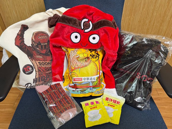 An assortment of merchandise laid out on a chair, including a tote bag featuring a race car driver with 'SOON' and 'SVM' logos, a red panda plush toy holding a Nissin Cup Noodles package, black clothing in plastic, a red card holder, and two yellow packages of egg waffles.
