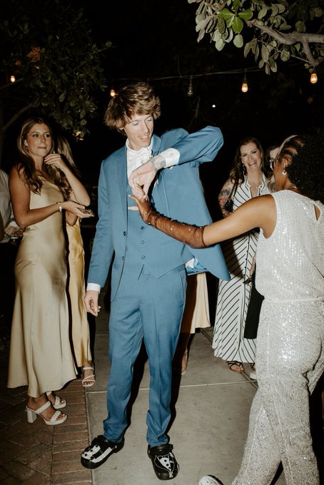 A young man in a light blue three-piece suit dances at an outdoor evening event with string lights, wearing sneakers, while other formally dressed guests watch and celebrate.