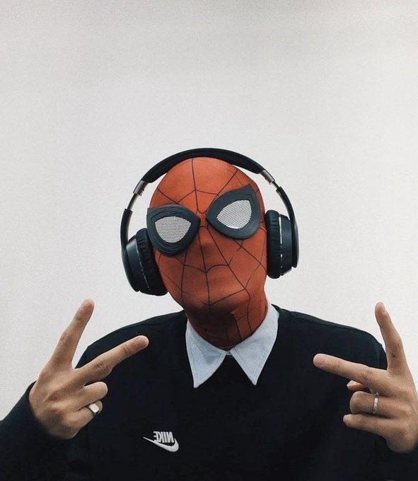 A person wearing a Spider-Man mask and a black Nike sweatshirt over a white collared shirt, wearing black headphones and making peace signs with both hands.