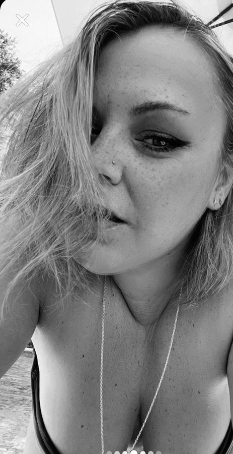 A black-and-white close-up selfie of a young woman with freckles and blonde hair blowing across her face, wearing a necklace.