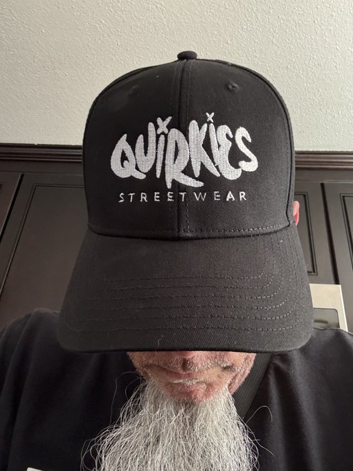 A man with a long gray beard wearing a black baseball cap with white text reading 'QUIRKIES STREETWEAR'.