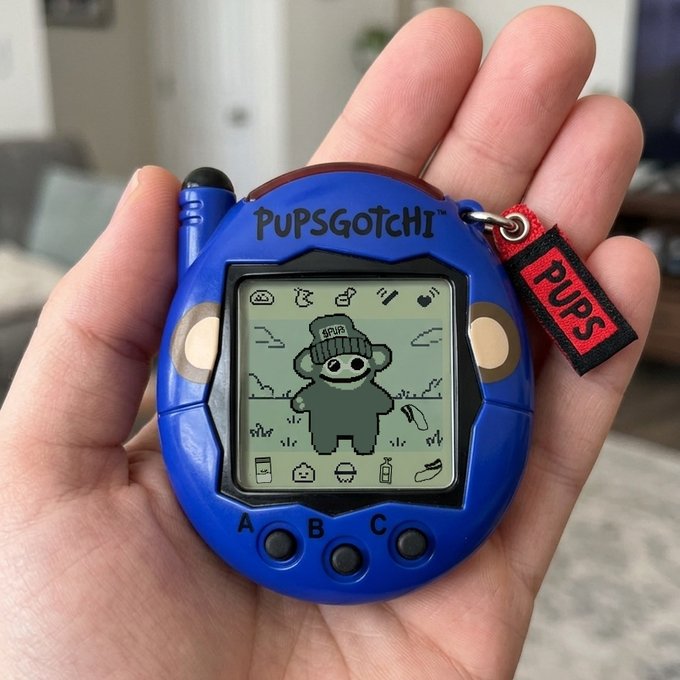 A hand holds a blue "PupsGotchi" virtual pet device featuring a pixelated dog character wearing a beanie, with a "PUPS" red tag attached. The post humorously references the challenge of keeping this digital puppy alive after losing it twice, hoping for success on the third attempt.