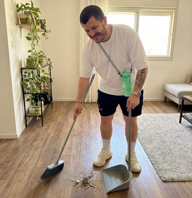 The image shows a man wearing white sneakers, holding a broom with a dustpan, cleaning up debris from the floor. The room appears to be a living space or bedroom, as there are potted plants placed around the area. A couch can also be seen in the background, suggesting that this is likely a residential setting.

The post about sweeping the floor seems to be related to the man's actions, possibly discussing his cleaning process or techniques. The image captures the essence of the post by showing the man actively engaged in cleaning up debris from the wooden floor using a broom and dustpan.