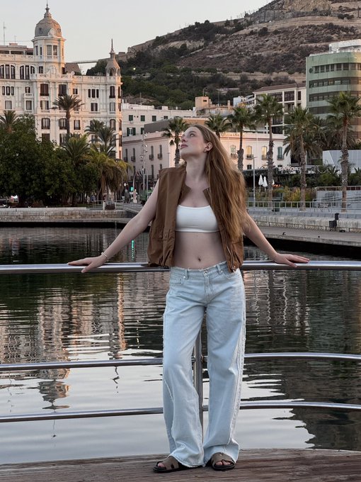 A young woman with long brown hair, wearing a brown vest over a white crop top and light blue jeans, poses by a railing overlooking a waterfront with buildings and palm trees in the background.