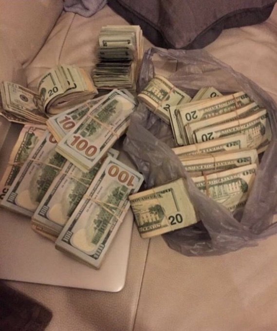 A photograph showing a large pile of banded US currency, including $20 and $100 bills, resting in and around a clear plastic bag on a bed.