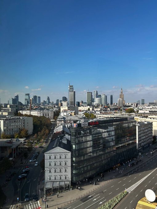 An elevated daytime view of a cityscape shows a skyline with several tall skyscrapers and the Palace of Culture and Science in the distance, with a modern glass building in the foreground.