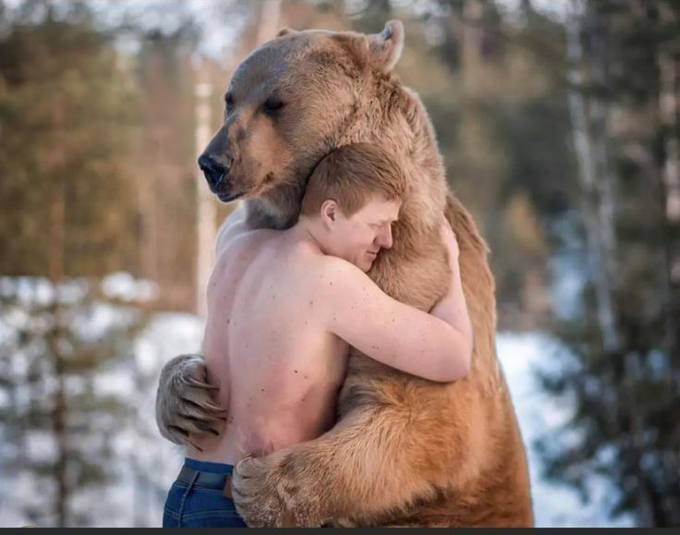A photoshopped or AI-generated image showing a shirtless young man hugging a large brown bear in a snowy forest setting.