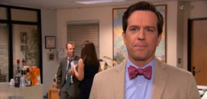 Ed Helms as Andy Bernard from The Office wearing a tan suit and a distinctive purple bow tie.