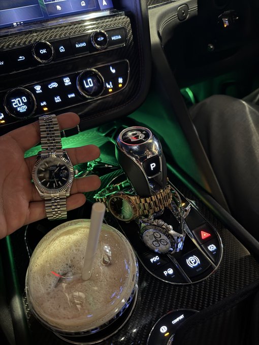 A nighttime photo inside a luxury vehicle featuring a hand holding a silver Rolex watch, with a gold watch and car keys resting on the center console next to a drink.