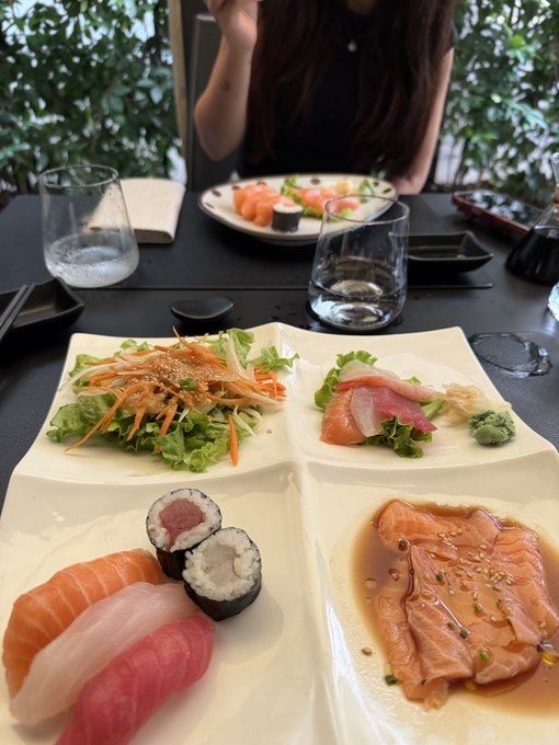 A plate of assorted sushi, sashimi, and salad served at a restaurant with another plate and a person visible in the background.