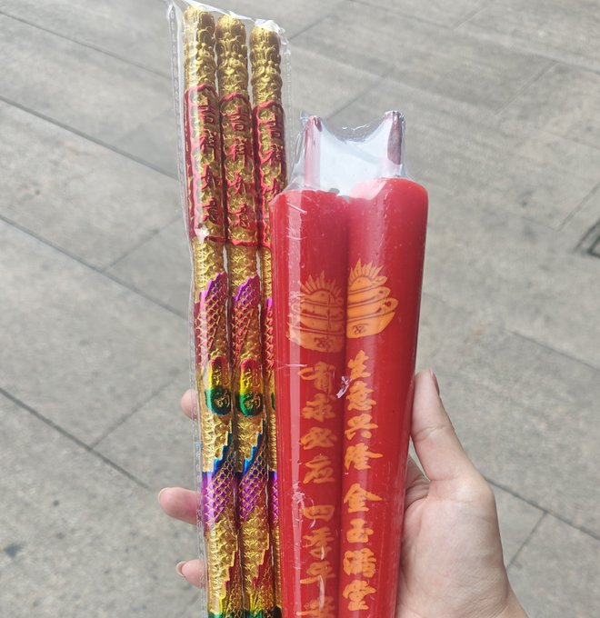 A hand holding red ceremonial candles and decorative sticks with Chinese characters and gold and pink patterns, wrapped in plastic packaging.