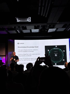 A man stands at a podium with a large screen behind him, giving a presentation or speech about Decentralized Knowledge Graphs (KG) during an event taking place in Ljubljana. The room appears packed, indicating a high interest and engagement level from attendees. The post on the image provides additional context by stating that it is a packed meeting of @cursor_ai at today's #cursor_ai meetup in Ljubljana with BranaRakic showing an update on Decentralized Knowledge Graph V10 live on stage, emphasizing the value and importance of this new concept.