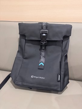 The image features a black backpack sitting on top of a couch, with the words "bitget" written on it. The backpack appears to be new and unused, suggesting that it has been recently purchased or received as a gift. The presence of the backpack on the couch indicates that someone is likely in their home or living space, possibly preparing for an outing or returning from one.