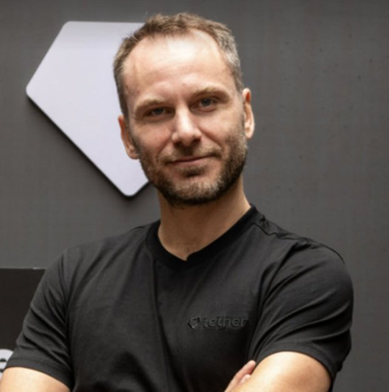 A man with short hair and stubble wearing a black t-shirt, posing with crossed arms in front of a gray wall with a white hexagonal decoration.