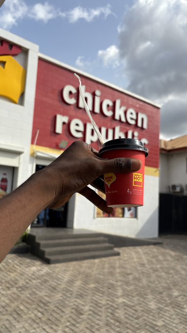 A first-person photo of a hand holding a red beverage cup with a straw in front of a 'Chicken Republic' fast food restaurant. The location appears to be in Africa based on the branding.