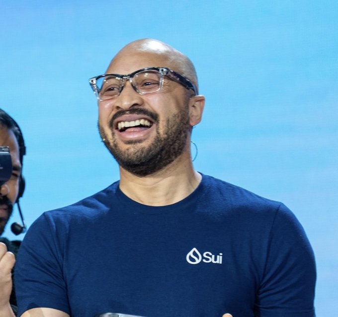 A smiling bald man with a beard and glasses wears a dark blue t-shirt featuring the white 'Sui' logo.