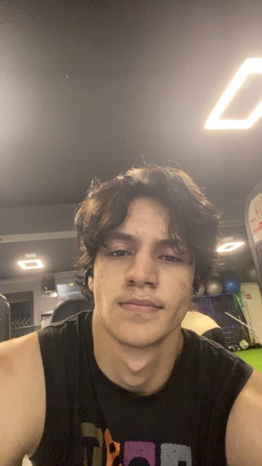A young man with dark wavy hair takes a gym selfie, wearing a black tank top with partially visible colorful text. The image aligns with the motivational post, capturing a quiet, determined moment that reflects the message of showing up and staying consistent with the work.