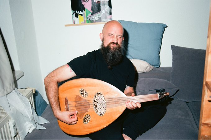 A bearded, bald man in a black t-shirt sits on a gray couch smiling as he holds a wooden oud (a Middle Eastern stringed instrument) with intricate rosettes.
