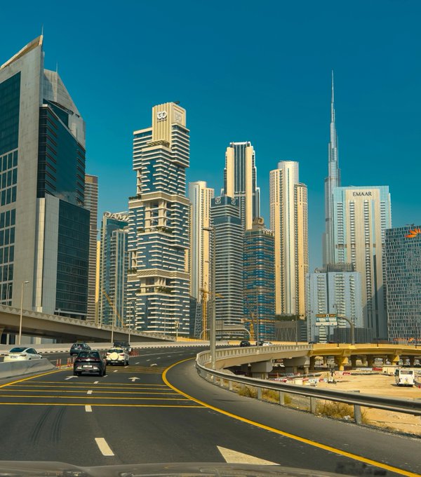 A photograph of a city skyline featuring modern skyscrapers, including the Burj Khalifa, with the EMAAR logo visible on a building, viewed from a curved highway under a clear blue sky.
