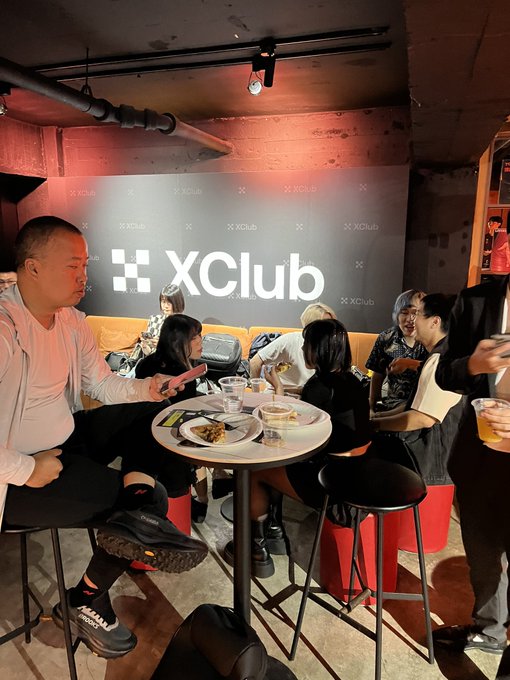 A group of people sit around a small table at the XClub venue, with the 'XClub' logo visible on a backdrop behind them and red lighting illuminating the brick-walled lounge area.