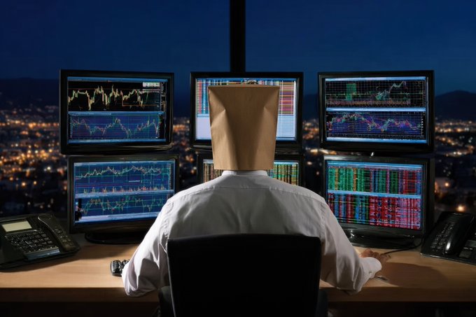 A person wearing a paper bag over their head sits at a desk facing six monitors displaying financial trading charts and data at night.