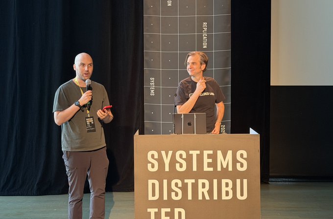 Two men present at the "Systems Distributed" conference, with one speaking into a microphone on the left and another standing behind a podium with an Apple laptop on the right, against a backdrop featuring a grid labeled "REPLICATION" and "SYSTEMS." This aligns with the introduction of Zooko, the creator of Zooko's Triangle and Zcash, being presented at the event.