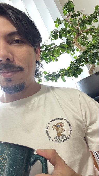 The image features a man wearing a white t-shirt that has an amusing cartoon of a monkey holding a cup, with the text "GM having coffee and listening in on Meet at the Clubhouse with the @BoredApeYC". The man is holding a green mug filled with coffee, which adds to the overall theme. The background includes a potted plant and a window, creating an indoor setting for this scene.