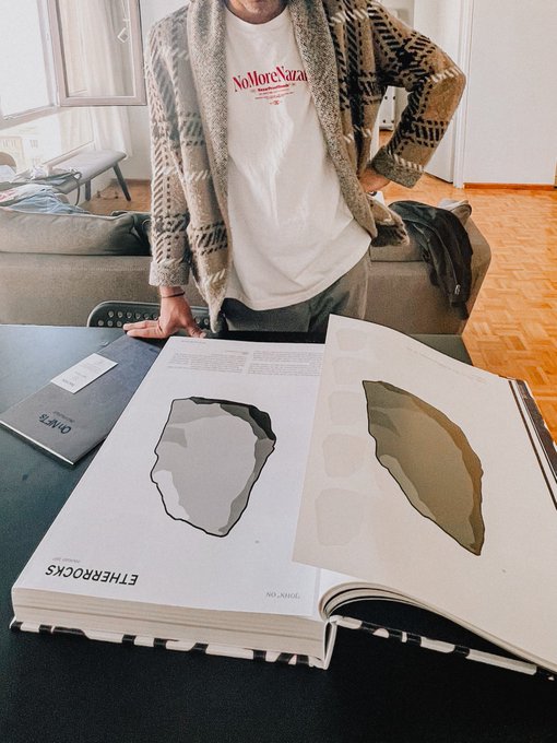 A person wearing a patterned cardigan and a white 'No More Nazaraj' t-shirt stands behind a table displaying a large book titled 'ETHEROCKS' open to illustrations of rocks or stones.