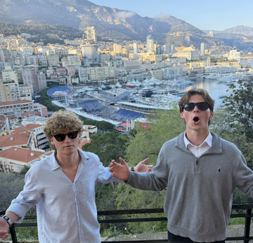 Two young men posing playfully in front of a scenic coastal city view with a harbor and mountains in the background.